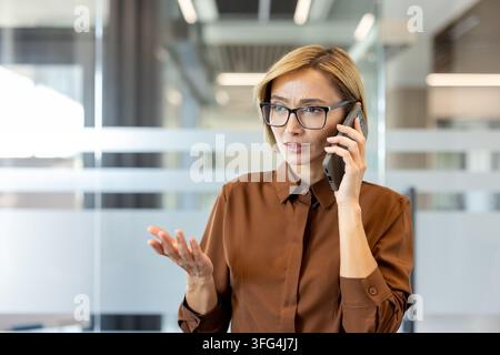 Donna d'affari su uno smartphone in un ufficio moderno, indossa occhiali e una camicia marrone, sembra arrabbiata e gesticola durante una chiamata di lavoro seria e stressante su sfide professionali Foto Stock