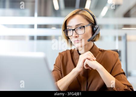 Donna d'affari sicura di sé con auricolare e occhiali, che lavora su un laptop in un ufficio, attivamente impegnata nel telemarketing, assistenza clienti e comunicazione online Foto Stock
