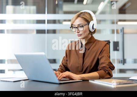 Donna d'affari concentrata sul suo lavoro, con occhiali e cuffie mentre digita su un laptop, dimostrando lavoro da remoto, concentrazione e produttività in un ambiente d'ufficio contemporaneo Foto Stock