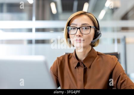 Rappresentante professionale del servizio clienti con auricolare e occhiali, seduta a una scrivania in un ufficio moderno, lavora e comunica con i clienti Foto Stock