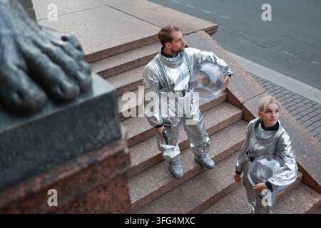 Ritratto di un uomo e una donna in tute spaziali argentate con caschi trasparenti. Due personaggi calmi di una storia retro-futuristica sui gradini di granito di un Foto Stock