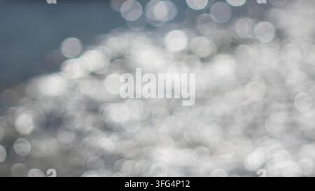 Sfondo astratto con numerosi cerchi di luce bokeh bianchi e grigi scintillanti che si riflettono su una superficie d'acqua blu-grigio morbida. Foto Stock