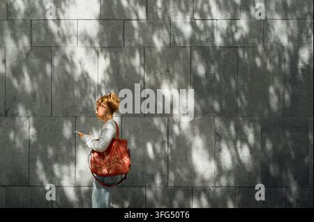 Giovane donna che usa lo smartphone mentre passa davanti a un muro di pietra Foto Stock