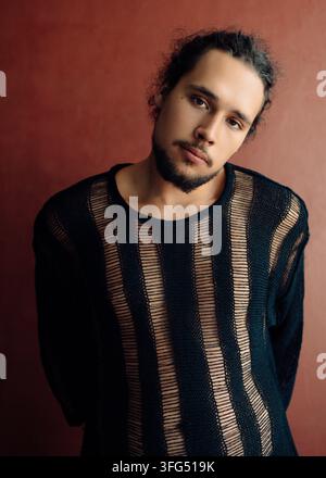 Giovane uomo barbuto in maglione a righe di lana davanti a uno sfondo rosso, espressione neutra e calma, ritratto in studio Foto Stock
