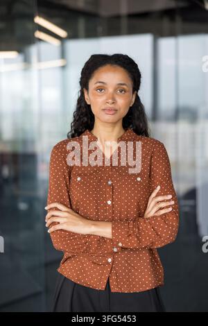 Giovane donna professionista in piedi con un'espressione seria e sicura, braccia incrociate, indossa una camicia a pois marroni, rappresentando la leadership e il successo in un ambiente aziendale contemporaneo Foto Stock