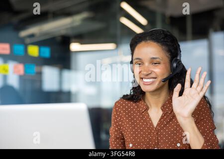 Donna d'affari sorridente con auricolare che partecipa a una videochiamata o riunione online amichevole dal suo ufficio moderno, salutando i partecipanti con un caloroso W Foto Stock