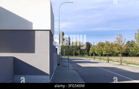 Facciata moderna grigia e bianca di un edificio con marciapiede in cemento, strada e una fila di piccoli alberi a destra. Sfondo per spazio di copia. Foto Stock