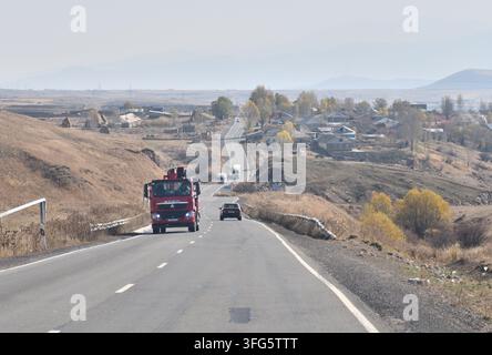 Un camion commerciale rosso percorre un'autostrada a due corsie asfaltata che attraversa un villaggio rurale in una regione montuosa dell'Armenia. Foto Stock
