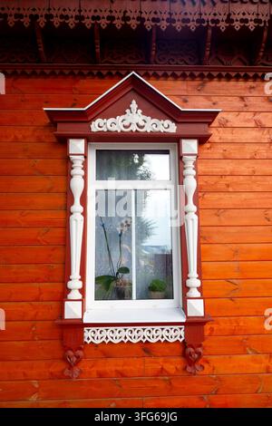 Vista dettagliata di una cornice di finestra in legno tradizionale russa ornata con intricate intagli decorativi bianchi e rossi su uno sfondo arancione brillante Foto Stock