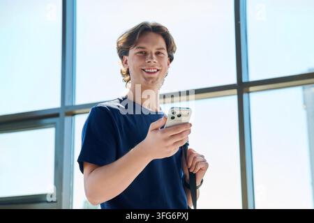 Giovane studente maschio che usa uno smartphone, in piedi vicino a una finestra nel campus e guarda verso la fotocamera Foto Stock