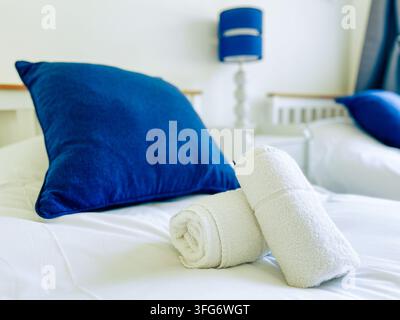 Asciugamani bianchi appena arrotolati disposti su un letto in una camera doppia di un hotel con cuscini blu navy. Foto Stock