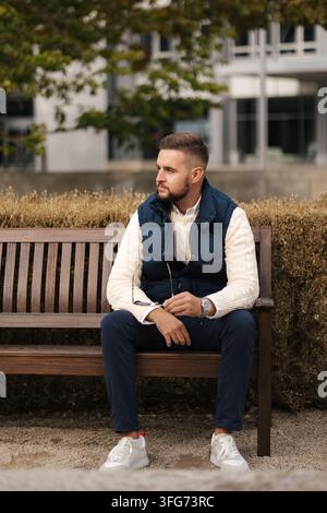 Prendersi un momento per rilassarsi all'aperto, un uomo vestito in modo casual si siede comodamente in un gilet elegante circondato dal fogliame autunnale. L'ambiente accogliente, naturale Foto Stock