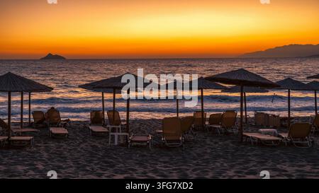 Tramonto sulla spiaggia di Kalamaki a Creta, Grecia, con ombrelloni di paglia e sdraio vuoti silouettati contro il cielo arancione luminoso sopra il Mar Libico Foto Stock