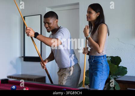 Uomo appoggiato, che stringe la stecca e prepara il tiro mentre una donna osserva le palle da biliardo su un tavolo bordeaux a casa Foto Stock