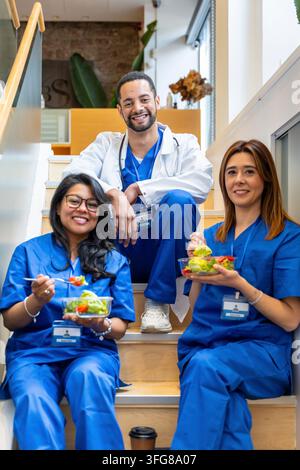 Tre professionisti sanitari diversi in camici – medico e infermiere – seduti sulle scale dell'ospedale, godendosi insalate fresche durante una pausa pranzo per promuovere Foto Stock