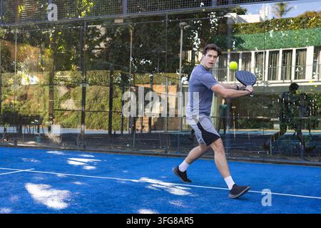 Uomo adulto che si lancia su un campo di paddle blu, tiene una pagaia, camicia grigia, fissa una palla gialla, spazio libero Foto Stock