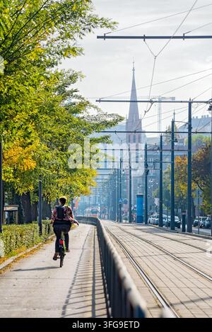 Ciclista che pedala lungo una pista ciclabile urbana accanto ai binari del tram a Budapest in una giornata autunnale soleggiata, con una guglia di chiesa in lontananza Foto Stock