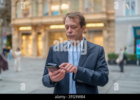 Un uomo d'affari di mezza età controlla il suo smartphone mentre è in piedi su una strada cittadina affollata. Foto Stock