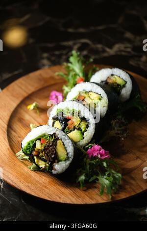 Rotoli di sushi ai vegetali freschi con avocado, cetriolo, verdure e riso su un piatto di legno, guarniti con erbe e fiori commestibili su un tavolo scuro. Foto Stock
