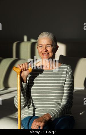 Donna anziana seduta su una panchina a casa, tiene un bastone di legno e mostra un anello indossando un top a righe Foto Stock