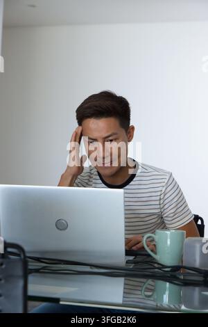 Uomo asiatico di mezza età seduto a casa, tocca la tempia, lavora su un laptop argentato con tazza e cavi Foto Stock