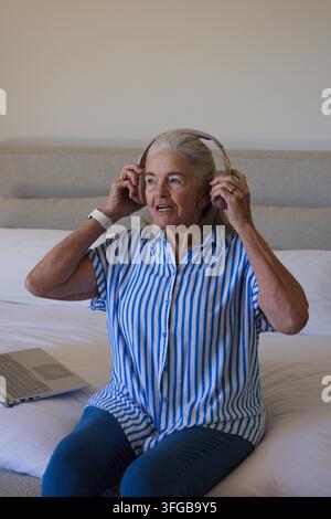 Donna anziana che regola delle cuffie sopra le orecchie mentre è seduta su un letto in camera da letto con un laptop nelle vicinanze Foto Stock