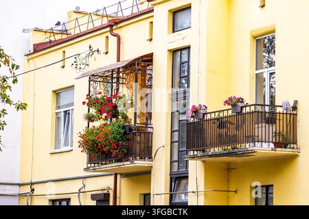 Vista esterna di un edificio residenziale giallo brillante con balconi in ferro battuto pieni di fiori in vaso rosso acceso e rosa alla luce del giorno. Clas Foto Stock
