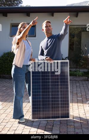Coppia diversa che tiene un pannello solare puntato verso il tetto su un vialetto in pietra fuori dalla casa in jeans Foto Stock