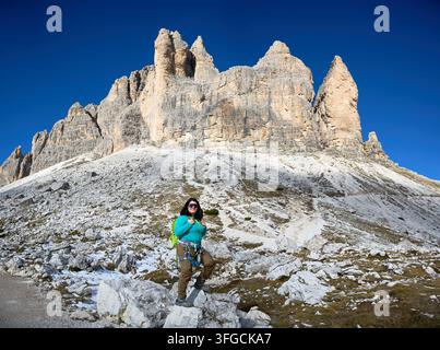 Arrampicatrice posa su pendice roccioso sotto le torri monumentali delle Dolomiti in Italia, indossa zaino, imbracatura via ferrata, illustrando fiducia, esplorazione alpina, fitness all'aperto, turismo d'avventura Foto Stock