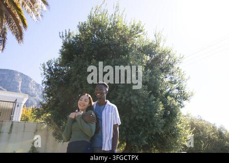 Coppia diversa in piedi che sorride davanti a una casa accanto a un basso muro di cemento e un cancello con la luce del sole Foto Stock