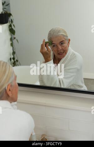 Donna anziana seduta alla toletta, indossa una vestaglia bianca, usa un pad konjac verde e guarda nello specchio Foto Stock