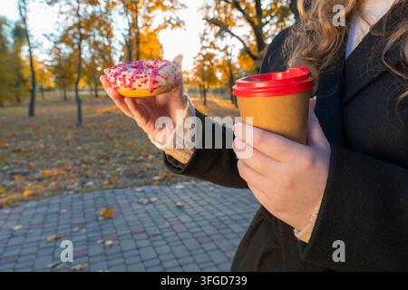 Primo piano di una donna in cappotto nero che tiene una tazza di carta per il caffè da asporto e una ciambella glassata con granella su uno sfondo autunnale del parco Foto Stock