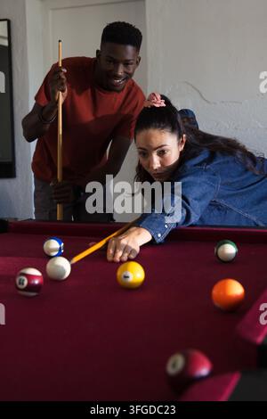 Amici diversi che giocano a biliardo con una donna che si china sul tavolo di feltro bordeaux allineando un tiro con le stecche Foto Stock