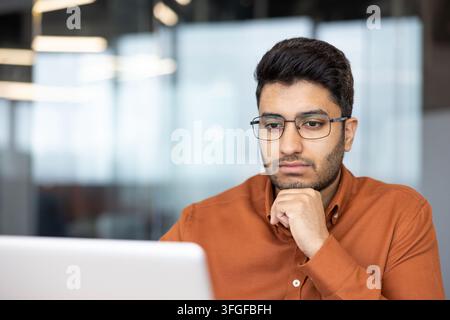 Giovane uomo con occhiali seduto davanti a un laptop in un ufficio moderno, con il mento appoggiato sulla mano mentre guarda lo schermo con un'espressione concentrata e pensierosa, incorniciato da una luce soffusa del giorno, sfondo vitreo sfocato, e Foto Stock