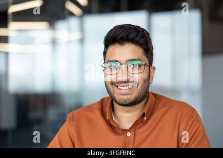 Ritratto di un uomo sudasiatico con occhiali e camicia arancione, che sorride alla telecamera all'interno di un ufficio moderno, luce naturale interna brillante, sfondo sfocato morbido, espressione amichevole, sicura di sé e accessibile Foto Stock