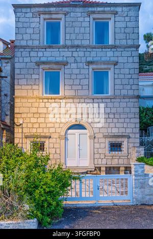 Facciata storica di una casa in pietra mediterranea con finestre blu e un cancello bianco al crepuscolo. La luce calma della sera evidenzia la trama della muratura, porta ad arco an Foto Stock