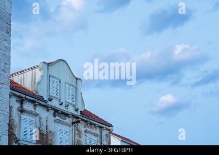 Edificio mediterraneo consumato con persiane in legno bianco e tetto di tegole in terracotta, che si staglia contro un cielo blu calmo con nuvole morbide, evocando una coa vintage Foto Stock