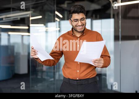Giovane professionista in camicia arancione con occhiali esamina documenti stampati in un moderno ufficio di vetro, tenendo carte mentre è in piedi alla luce naturale brillante per l'analisi e la pianificazione aziendale Foto Stock