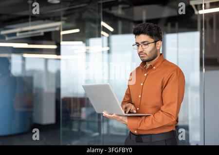 Giovane professionista in piedi in un ufficio moderno in vetro, che digita su un laptop mentre sembra concentrato e composto. Dettagli interni contemporanei luminosi evidenziano una giornata lavorativa produttiva guidata dall'efficienza professionale Foto Stock