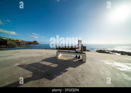 Una donna di 60-70 anni è seduta su una panchina, lungomare sulla costa meridionale con l'Oceano Atlantico alle spalle, Madera, Portogallo Foto Stock