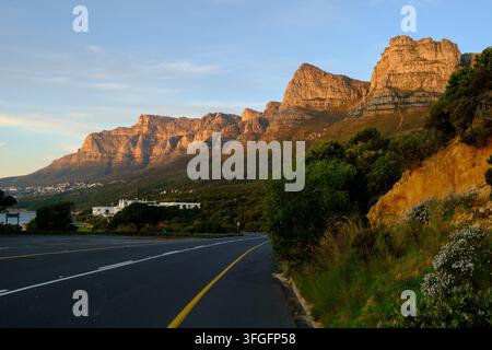 Un tramonto dorato bagna le Twelve Apostles e la catena del Table Mountain accanto a una strada costiera vicino al Capo, con scogliere frastagliate, vegetazione costiera e tra Foto Stock