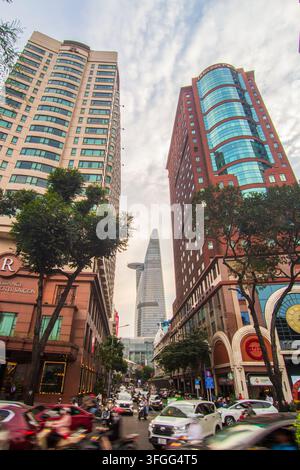 Traffico intenso e moto riempiono una strada fiancheggiata da alberi circondata da grattacieli moderni sotto un cielo nuvoloso a Ho Chi Minh City, Vietnam. Foto Stock