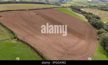 Vista aerea con drone di un trattore che ara un grande campo di terreno marrone ricco a Lanreath, Cornovaglia, Regno Unito. I campi adiacenti sono coperti di erba verde rigogliosa. Foto Stock