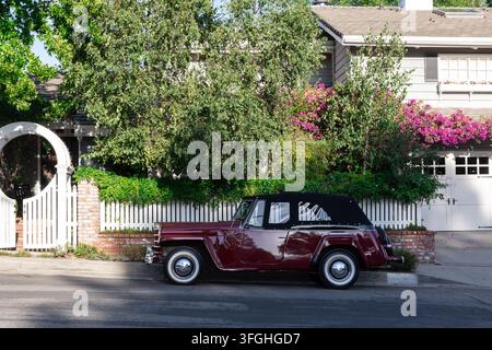 Los Angeles, California, USA - 12 settembre 2026: Auto bordeaux vintage parcheggiata lungo una strada residenziale accanto a una casa paesaggistica, recinto bianco, muro di mattoni Foto Stock