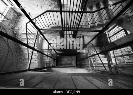 Guardando in basso in un pozzo dell'ascensore a gabbia abbandonato con rete metallica e scala circolare all'interno del sanatorio storico di Grabowsee in Germania. Foto Stock