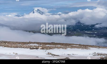 Paesaggio panoramico del Kamchatka con un maestoso vulcano innevato che si erge drammaticamente sopra un vasto mare di nuvole, circondato da picchi vulcanici frastagliati Foto Stock