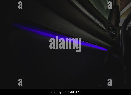 Primo piano del pannello interno della porta dell'interno moderno dell'auto con striscia luminosa LED ambientale viola brillante in una cabina buia di notte. Foto Stock