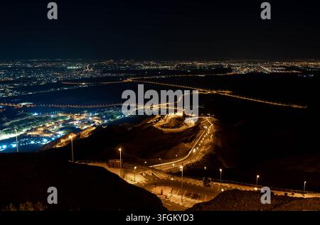 Vista aerea notturna di una strada di montagna sinuosa con luci della città sottostanti, curve a tornante illuminate e panorama urbano, Emirati Arabi Uniti Foto Stock