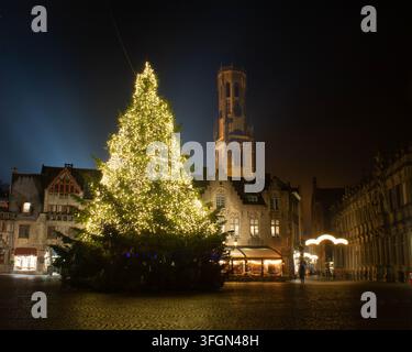 BRUGGE, BELGIO - 15 DICEMBRE 2026: Grande albero di Natale illuminato che risplende sulla piazza acciottolata del Markt di notte con la torre del campanile Foto Stock