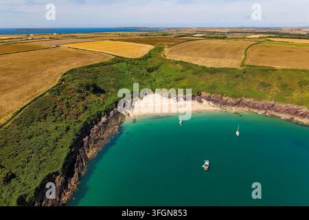 Paesaggio aereo di una baia sabbiosa circondata da scogliere e campi, tempo estivo soleggiato su acque costiere calme e barche a vela ormeggiate Foto Stock
